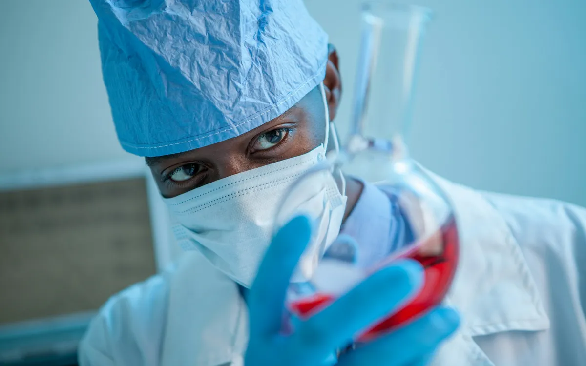 Profissional de laboratório com touca e máscara segurando um frasco com líquido vermelho