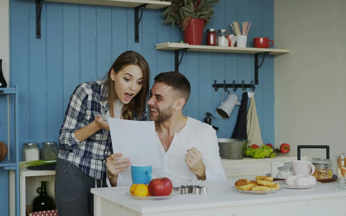 Casal sorrindo surpreso ao ler um documento na mesa da cozinha